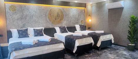 A hotel room with three beds, a large decorative wall, a modern ceiling light, and a potted plant.