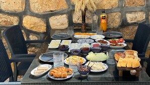 Ein Tisch ist reichhaltig mit Speisen gedeckt, darunter Brot, Käse und Obst, in einer Außenanlage mit Steinmauern.