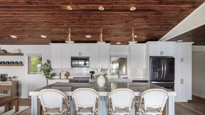 Une cuisine moderne avec des meubles blancs, un grand îlot central doté de tabourets, des appareils en acier inoxydable et un plafond en bois.