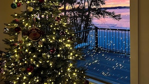 Un sapin de Noël décoré de guirlandes lumineuses et d’ornements, une pancarte indiquant « Lac Alexander », et une vue sur un coucher de soleil se reflétant sur l’eau.