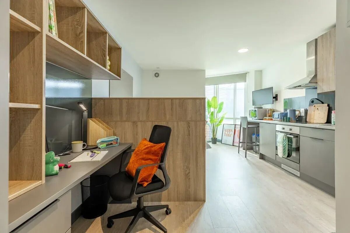 A modern kitchen with built-in appliances, a dining area with a black chair and orange cushion, and a workspace with a desk and shelves.