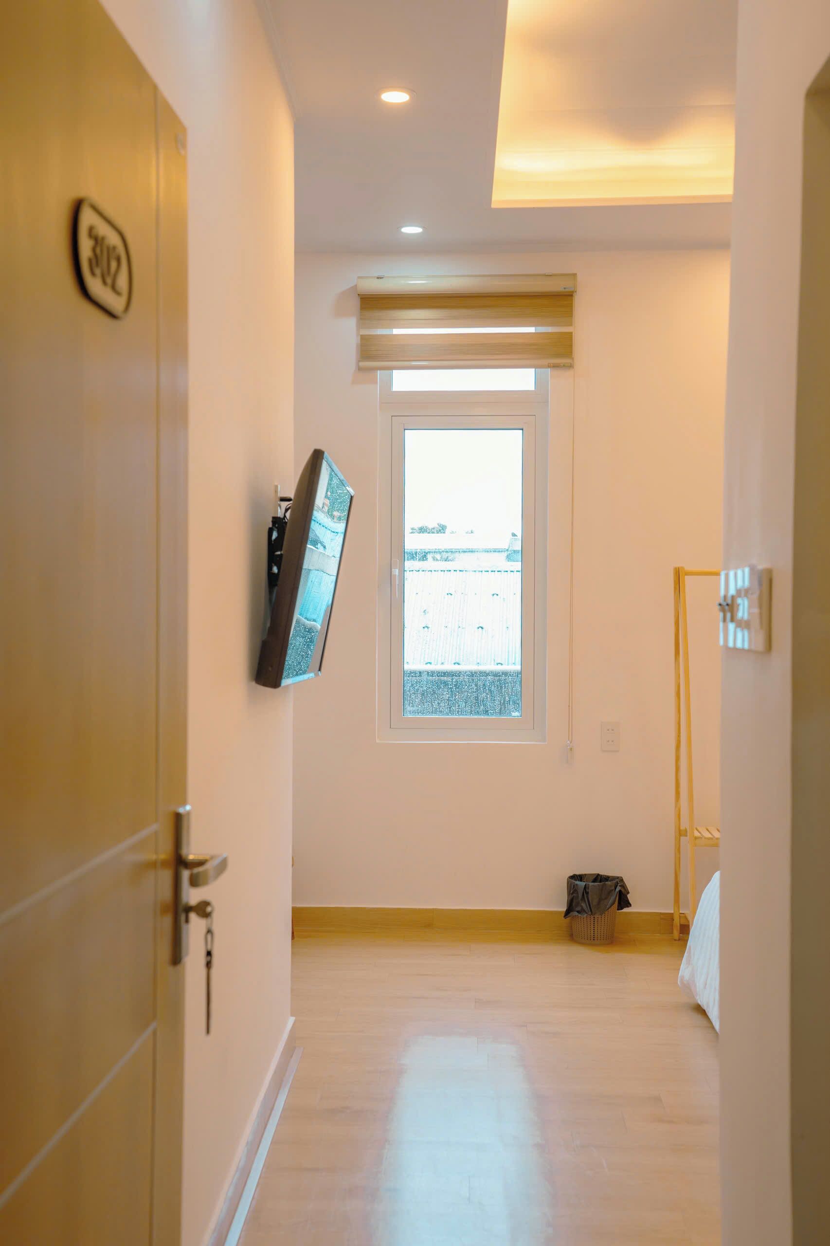 Una habitación de hotel con piso de madera, una ventana con persianas, un televisor montado en la pared y una puerta con placa de número.