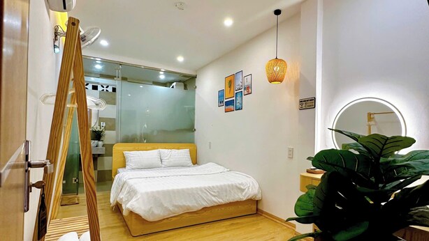 A modern hotel room with a wooden floor, a bed with white bedding, a standing fan, a large potted plant, and a mirror with a circular light. - Tom's Homestay Hue (Hue)