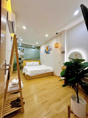 A modern hotel room with a wooden floor, a bed with white bedding, a standing fan, a large potted plant, and a mirror with a circular light. - Tom's Homestay Hue (Hue)