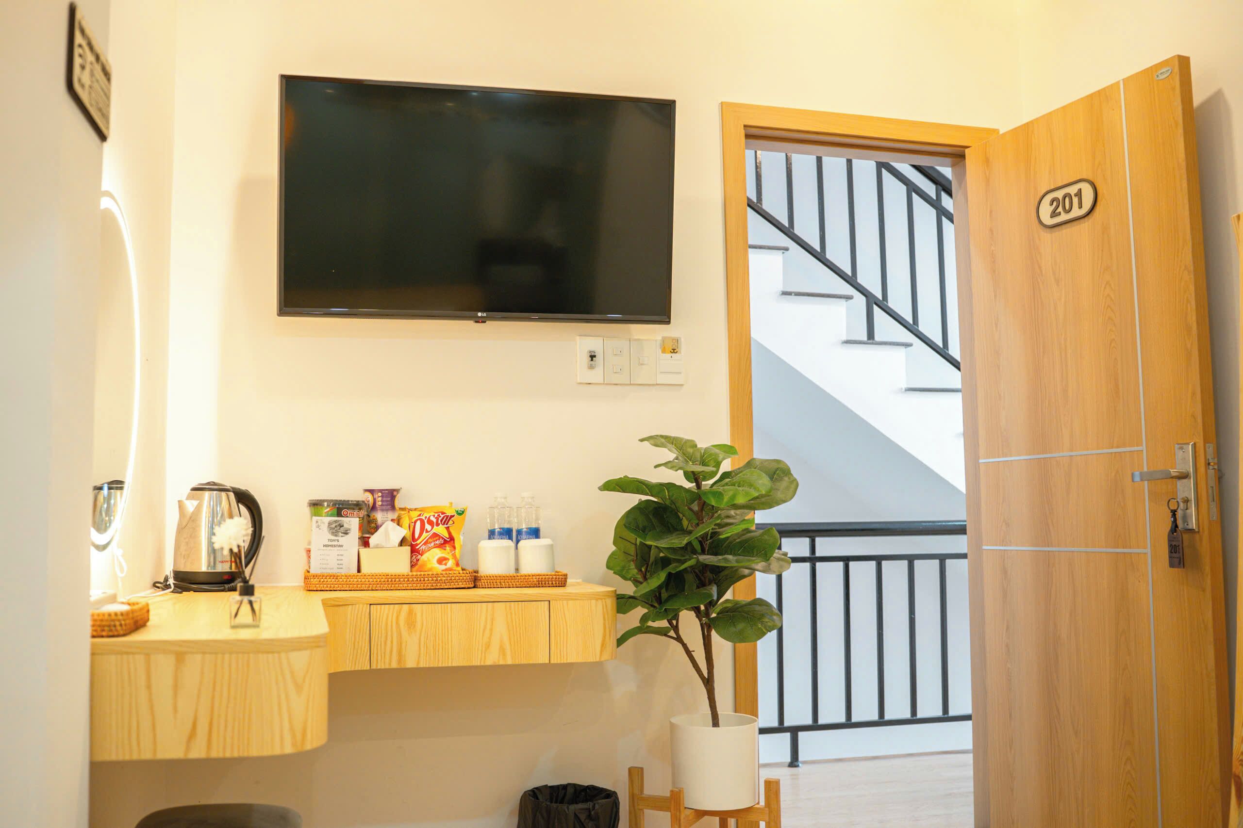 A hotel room with a TV mounted on the wall, a wooden counter with a kettle, snacks, and a potted plant.