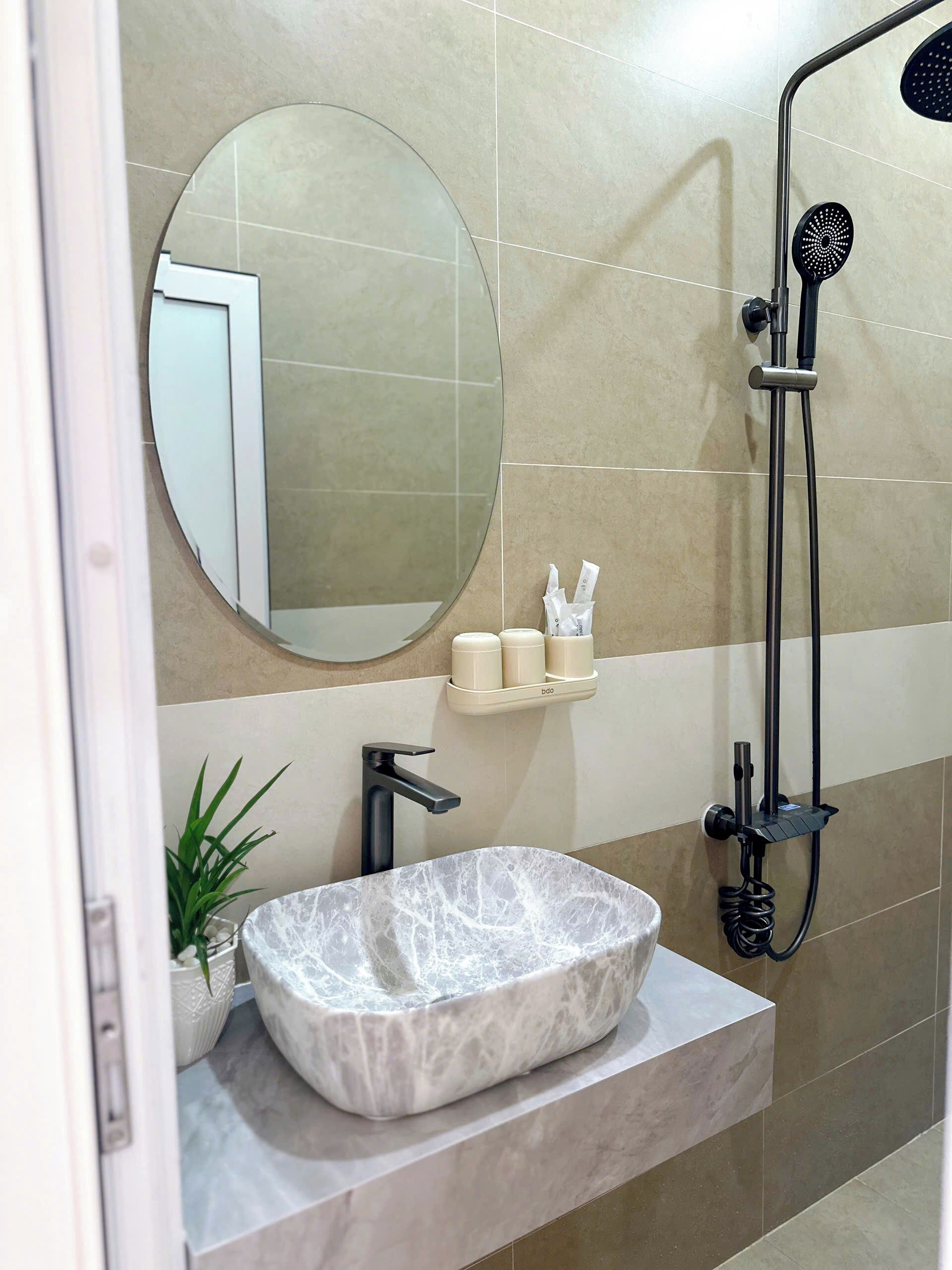 A bathroom with a marble sink, a round mirror, a handheld showerhead, and a shelf with toiletries.