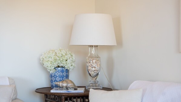 A room with a round wooden table, a white lamp, a vase with white flowers, and a decorative shell.