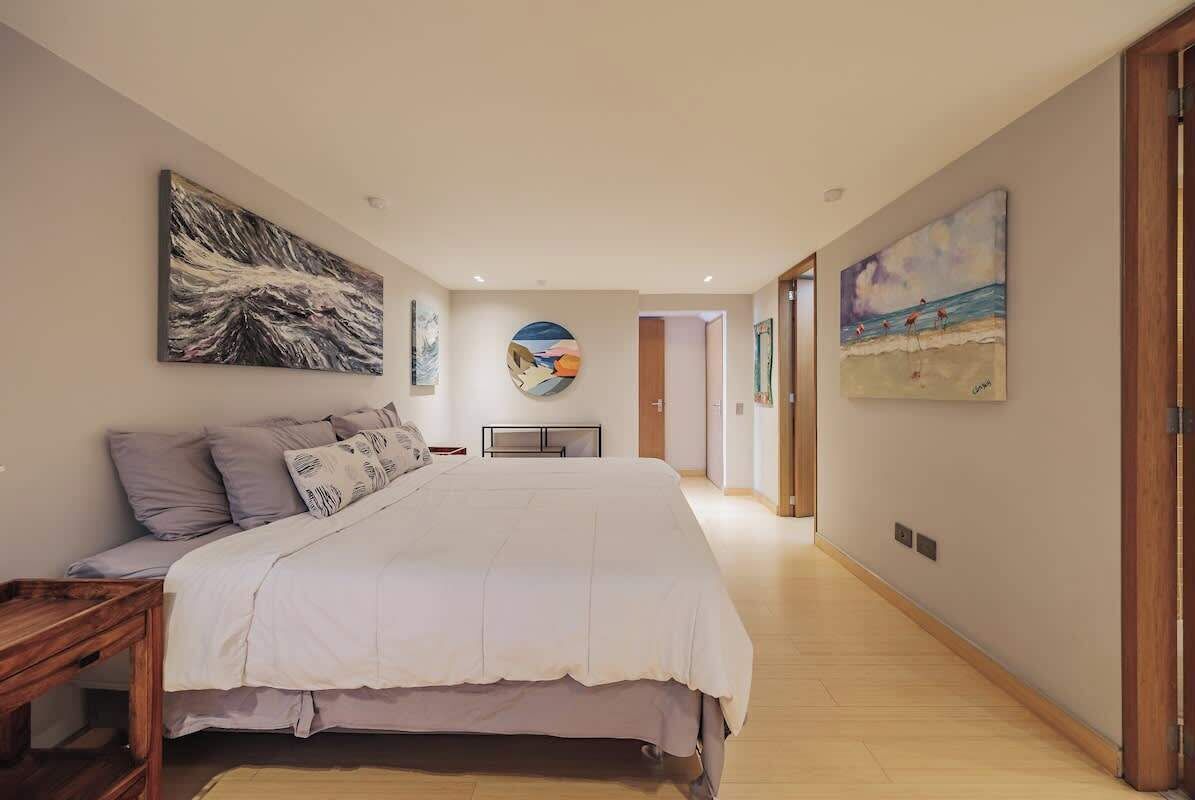 A bedroom with a large bed, a bedside table, and a wooden bench. There are three paintings on the walls.
