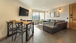A modern living room with a sofa, a dining table with chairs, a flat-screen TV mounted on the wall, and a large window providing a view of the outdoors.