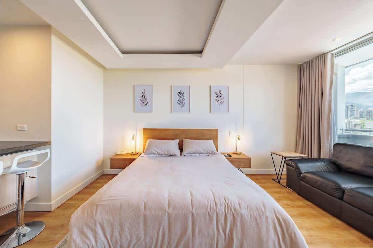 A modern hotel room with a large bed, a desk, and a leather sofa. There are three framed artworks on the wall and a city view through the window.
