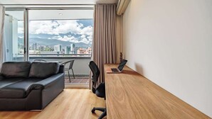 Une chambre moderne avec un canapé, un bureau en bois et un ordinateur portable. Une grande fenêtre offre une vue sur le paysage urbain et les montagnes.