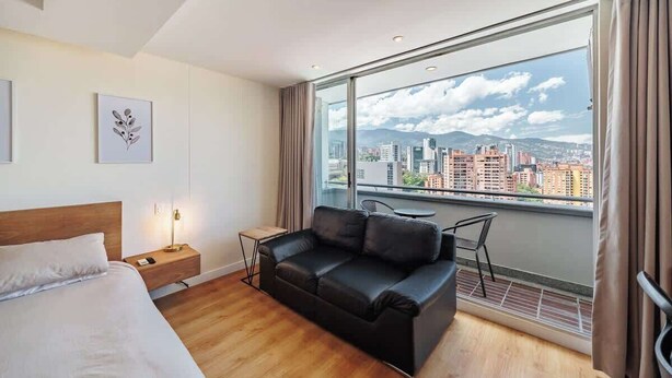 Une chambre d’hôtel moderne dotée d’une grande fenêtre, d’un canapé en cuir noir, d’un plancher en bois et d’un lit avec une literie blanche.