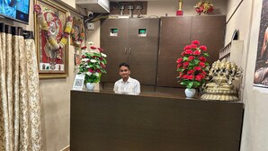 A reception area with a wooden reception desk, two potted plants with red flowers, a framed religious painting, and a security camera on the wall. - Hotel Nawanagar Residency (Jamnagar)