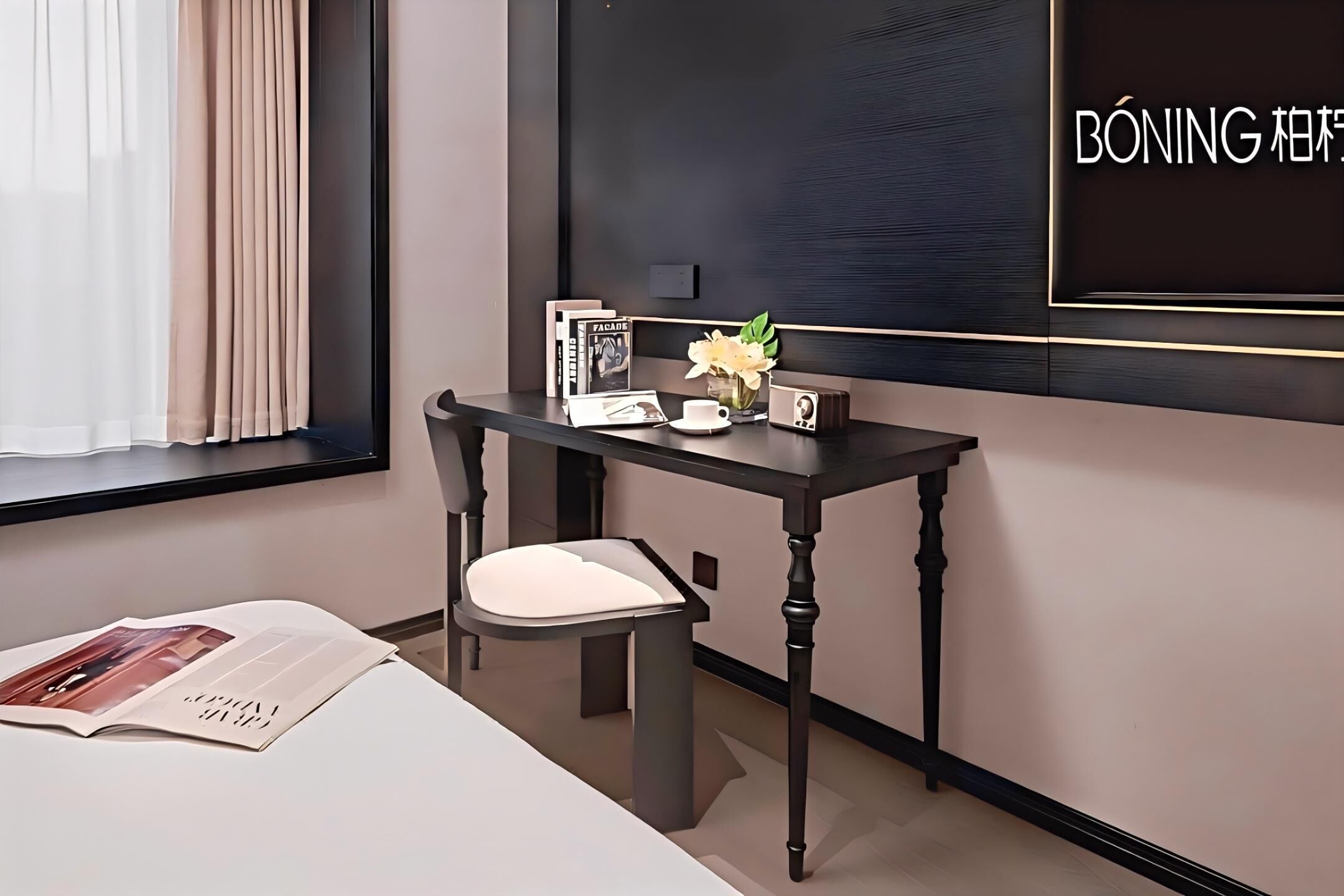 Ein modernes Hotelzimmer mit einem dunklen Holzbüro, einem weißen Sitzsessel, einem kleinen Tisch mit Blumen und einer Zeitschrift auf dem Bett.