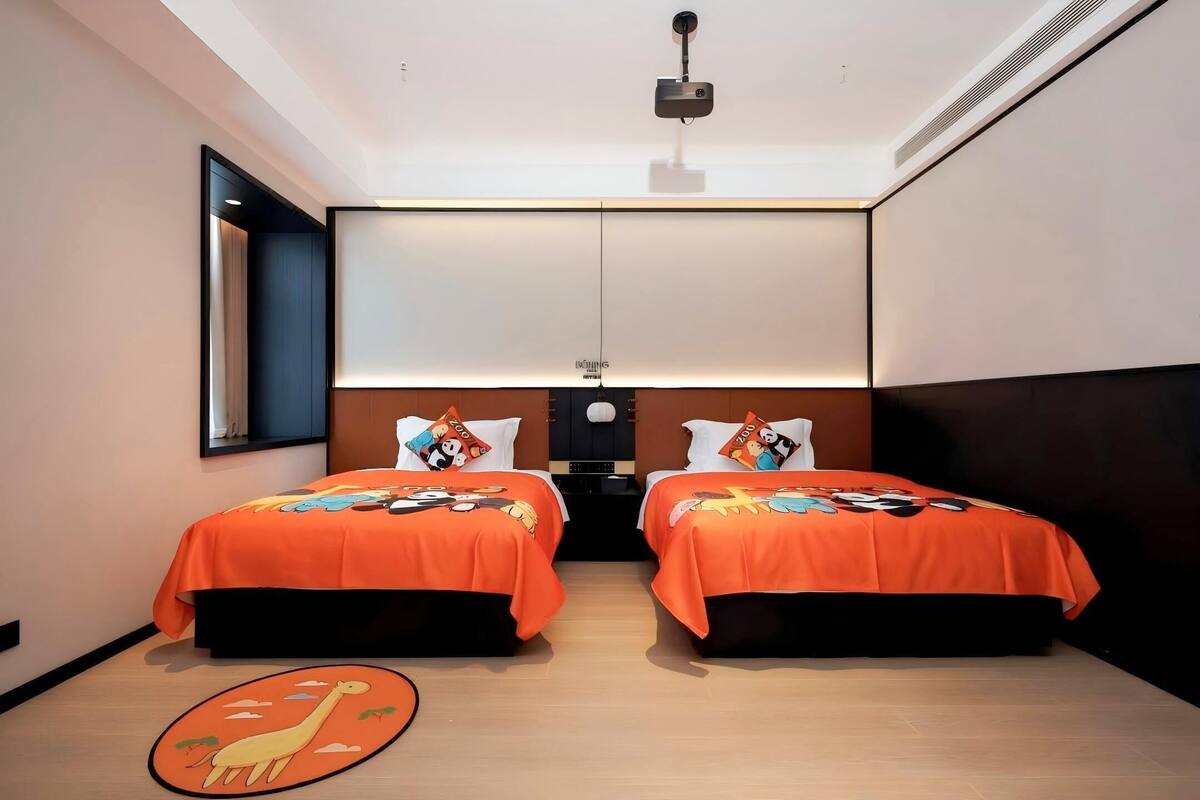 A hotel room with two beds, each with an orange bedspread and matching pillows, a ceiling-mounted projector, and a wooden floor.