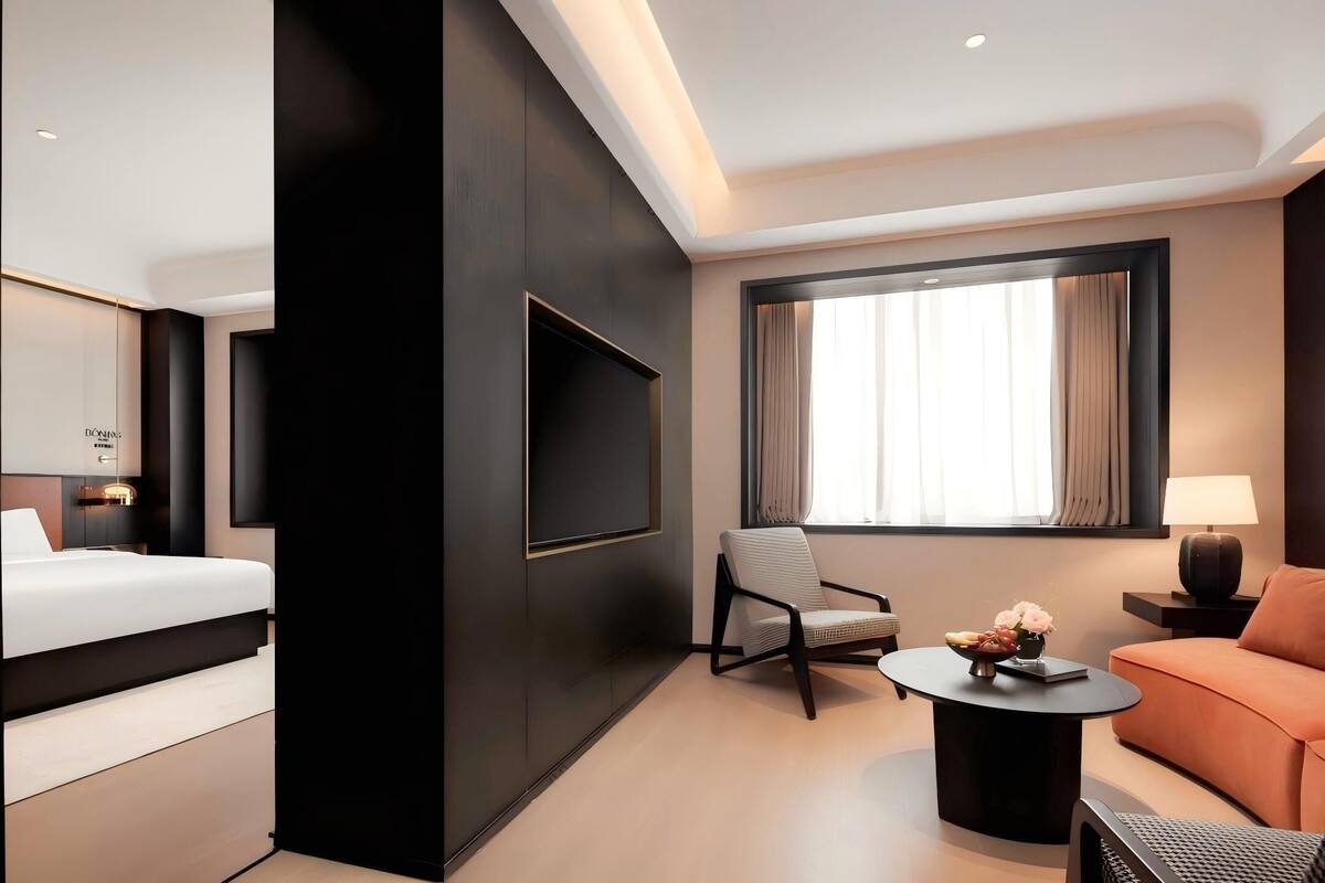 A modern hotel room with a bed, a flat-screen TV mounted on the wall, a seating area with a sofa and armchairs, a small round table with a vase of flowers, and a window with curtains.