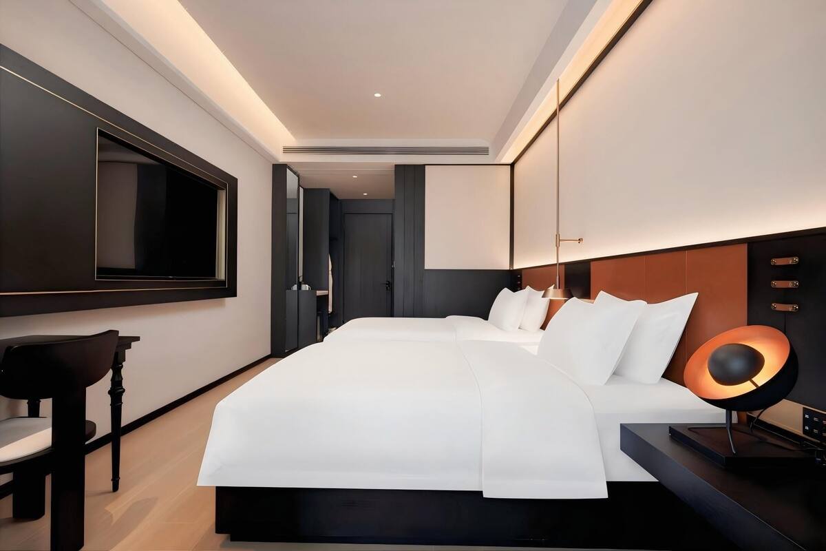 A modern hotel room with a large bed, a TV mounted on the wall, and a desk with a lamp.