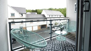 A balcony with two green chairs and a small table, overlooking a street with parked cars and houses.