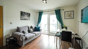 A modern living room with a grey sofa, a small side table, a dining area with a table and chairs, and a large window with curtains.
