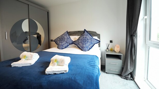 A neatly made bed with blue bedding and decorative pillows, a bedside table with a salt lamp, and a large mirror on the wardrobe door.