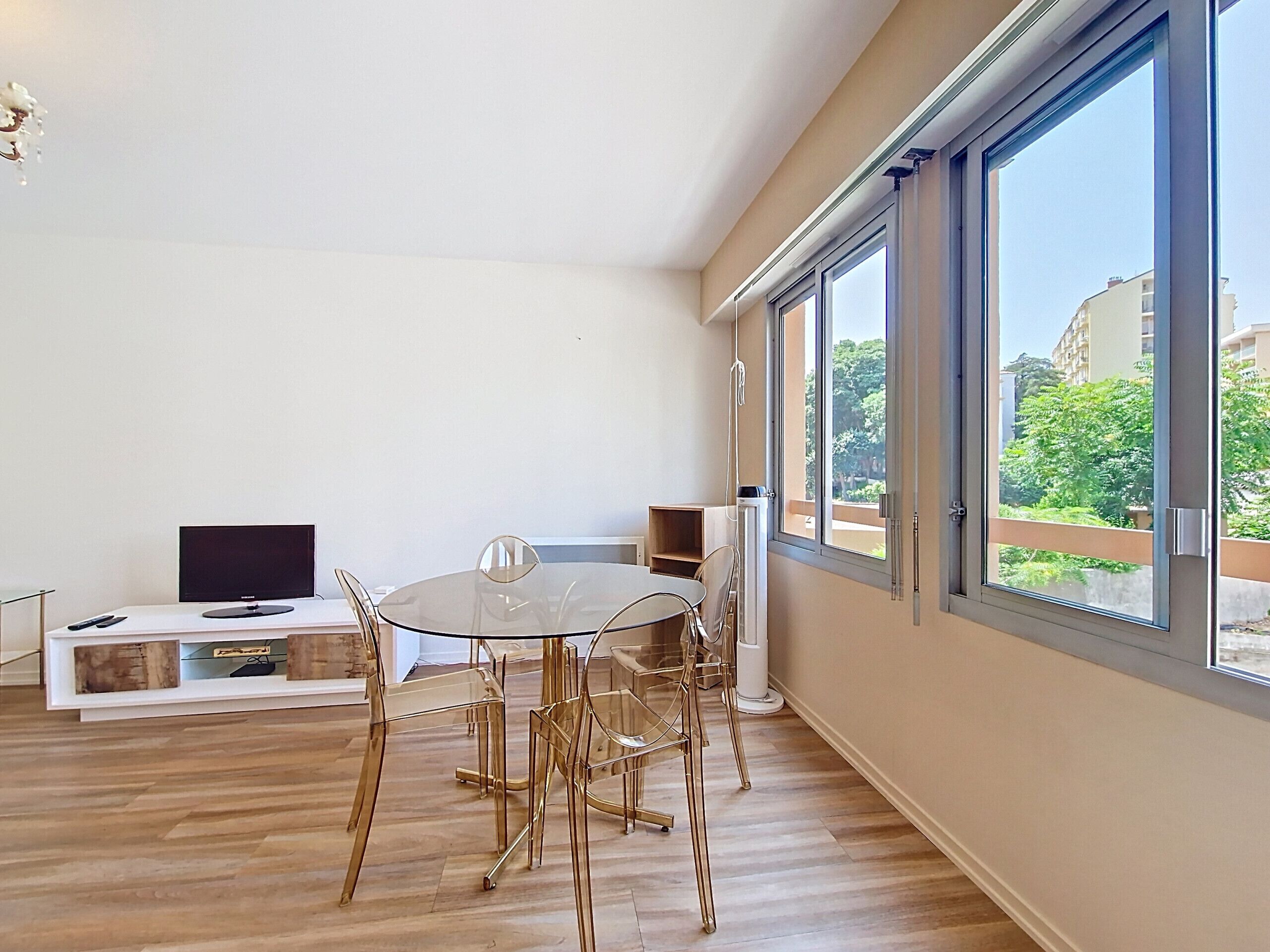 A modern living room with a glass table and chairs, a flat-screen TV, and a large window offering a view of buildings and trees.