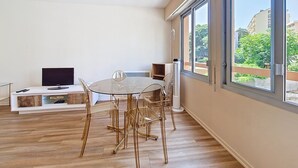 Una sala de estar moderna con una mesa y sillas de vidrio, un televisor de pantalla plana y un ventanal con vistas a edificios y árboles.