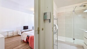 Habitación de hotel con cama, escritorio, televisión, ducha y puerta de vidrio que da al baño.
