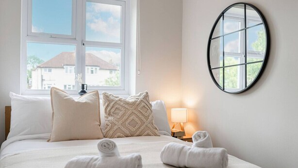 Ein ordentlich bezogenes Bett mit weißen Leinentüchern und zwei aufgerollten Handtüchern, eine Nachttischlampe und Blick auf ein Haus durch ein Fenster.