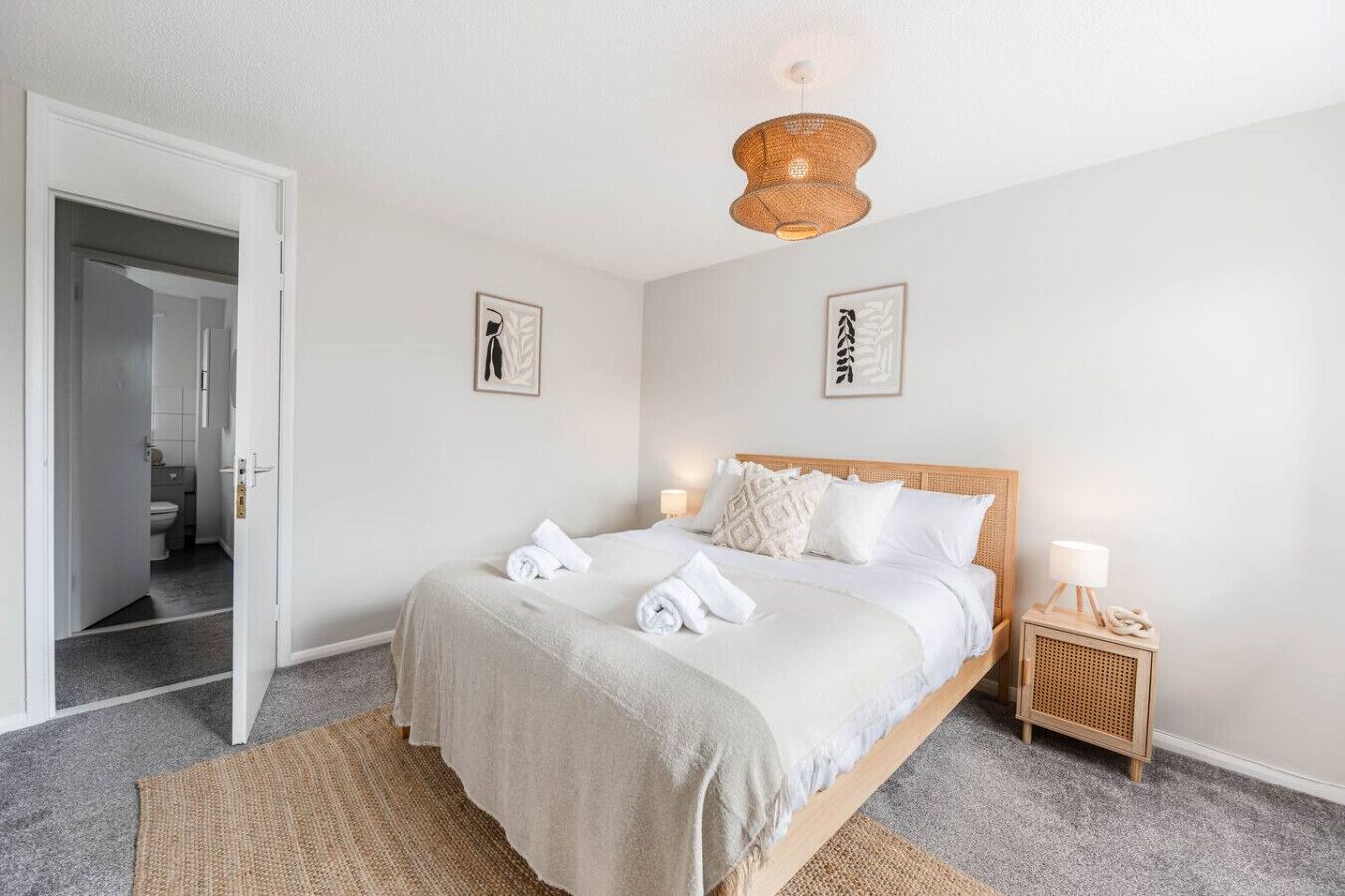 Ein Hotelzimmer mit einem Bett, einem Nachttisch mit Lampe, einer Tür zum Badezimmer und zwei an der Wand hängenden Kunstwerken.