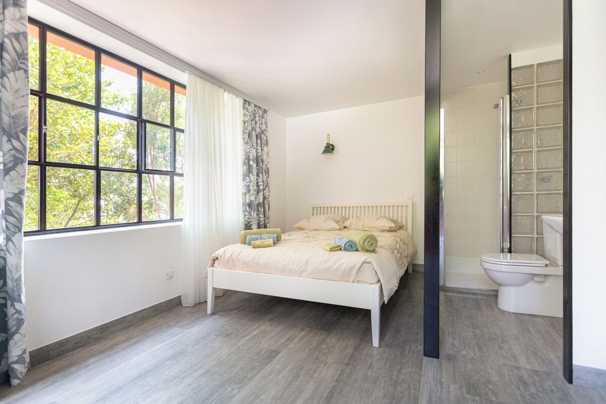 Quarto com uma janela grande, uma cama, uma mesa de cabeceira e um banheiro com vaso sanitário e chuveiro.