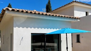 Un patio con mesa y sillas, una sombrilla azul y un ventanal de vidrio que da a un espacio interior.