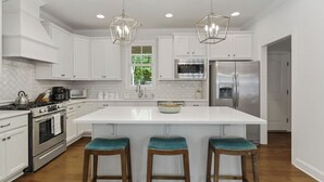 Una cocina moderna con armarios blancos, electrodomésticos de acero inoxidable, una isla central con taburetes y luminarias colgantes.