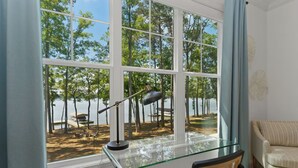 Una habitación con un escritorio de vidrio, una silla, una lámpara y vistas a una playa con árboles y un barco.