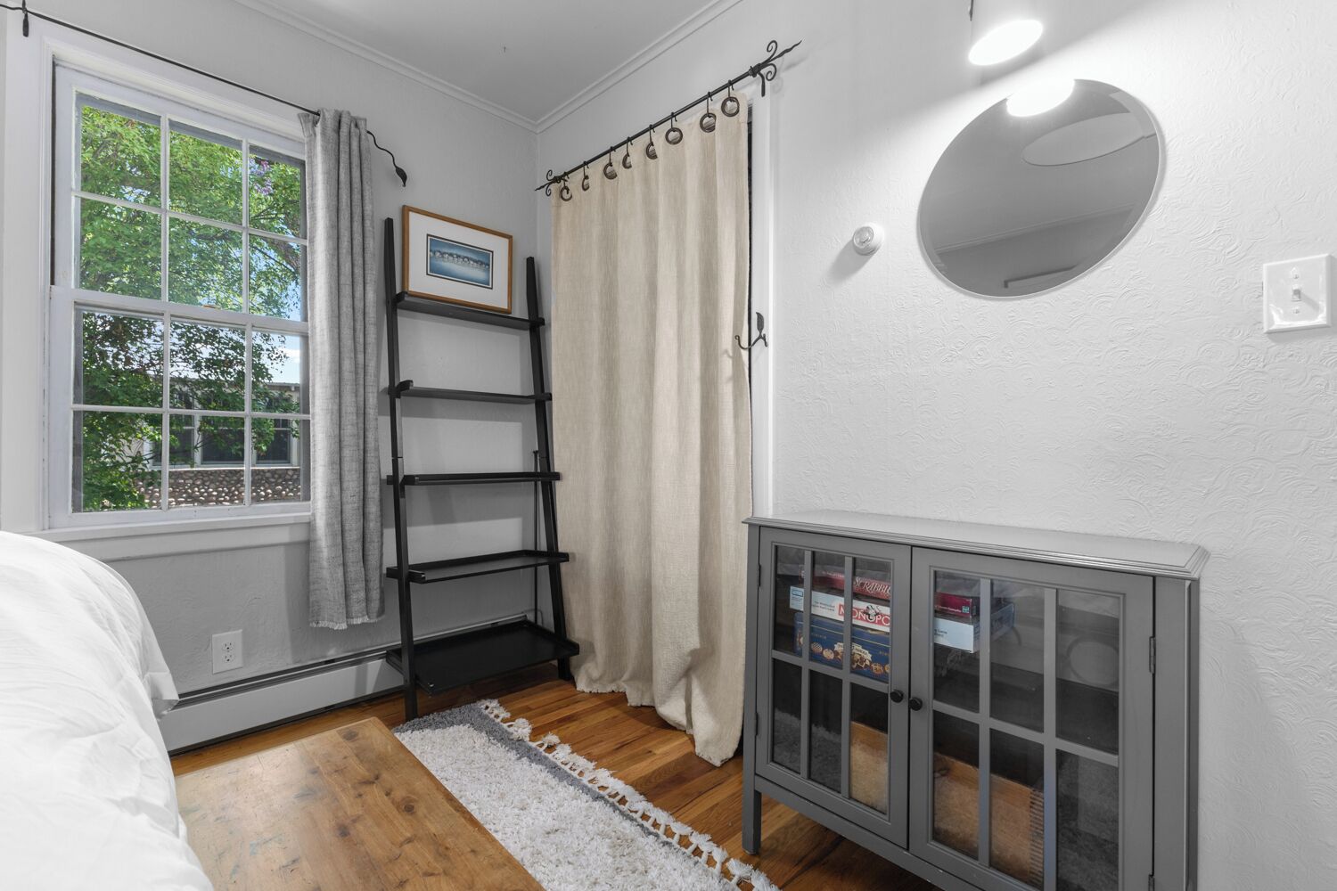 A bedroom with a bed, a window with curtains, a shelf, a radiator, and a framed picture on the wall.