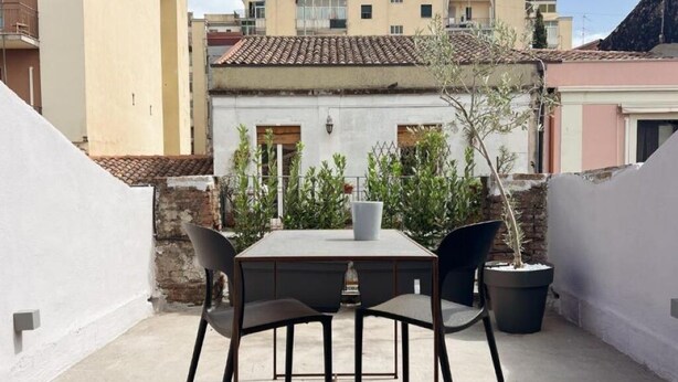 Une petite terrasse extérieure avec une table et des chaises, entourée de plantes en pot et offrant une vue sur les bâtiments voisins.