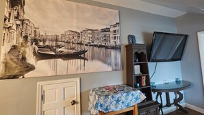 Une pièce décorée d’un grand tableau représentant un paysage urbain, avec un téléviseur fixé au mur, une petite table recouverte d’une nappe à motifs et une bibliothèque.