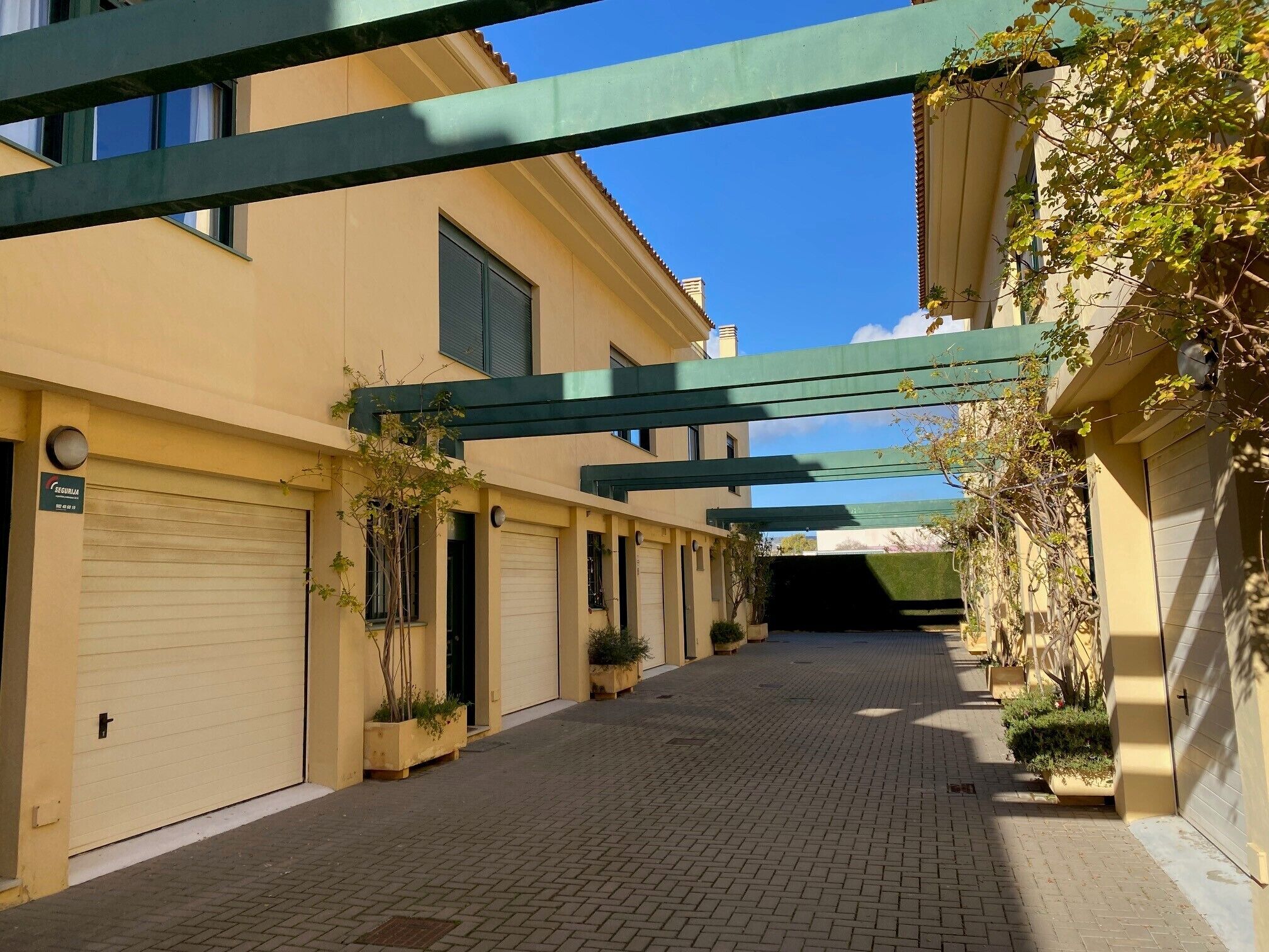 A paved alleyway with a row of closed garage doors on one side and a building with a green metal structure overhead.