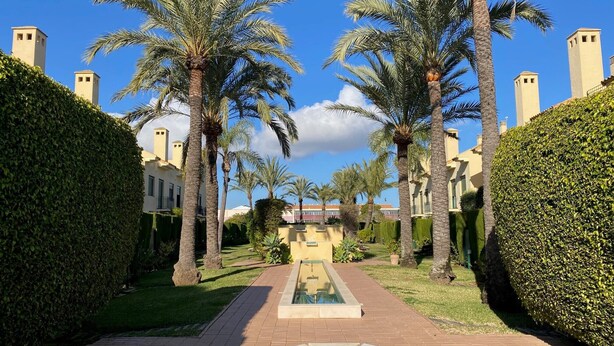 A pool area with palm trees and a hedge, surrounded by buildings with towers. - Luxury property in Javea Arenal with garden, roof terrace and shared pool (Xàbia)