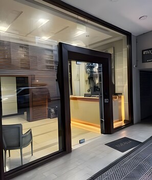 A modern hotel entrance with a glass door, a reception desk inside, and a chair outside. - collection R 1 (Santiago)