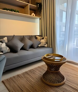 A modern living room with a grey sofa, patterned cushions, a woven side table, and a large window with curtains.