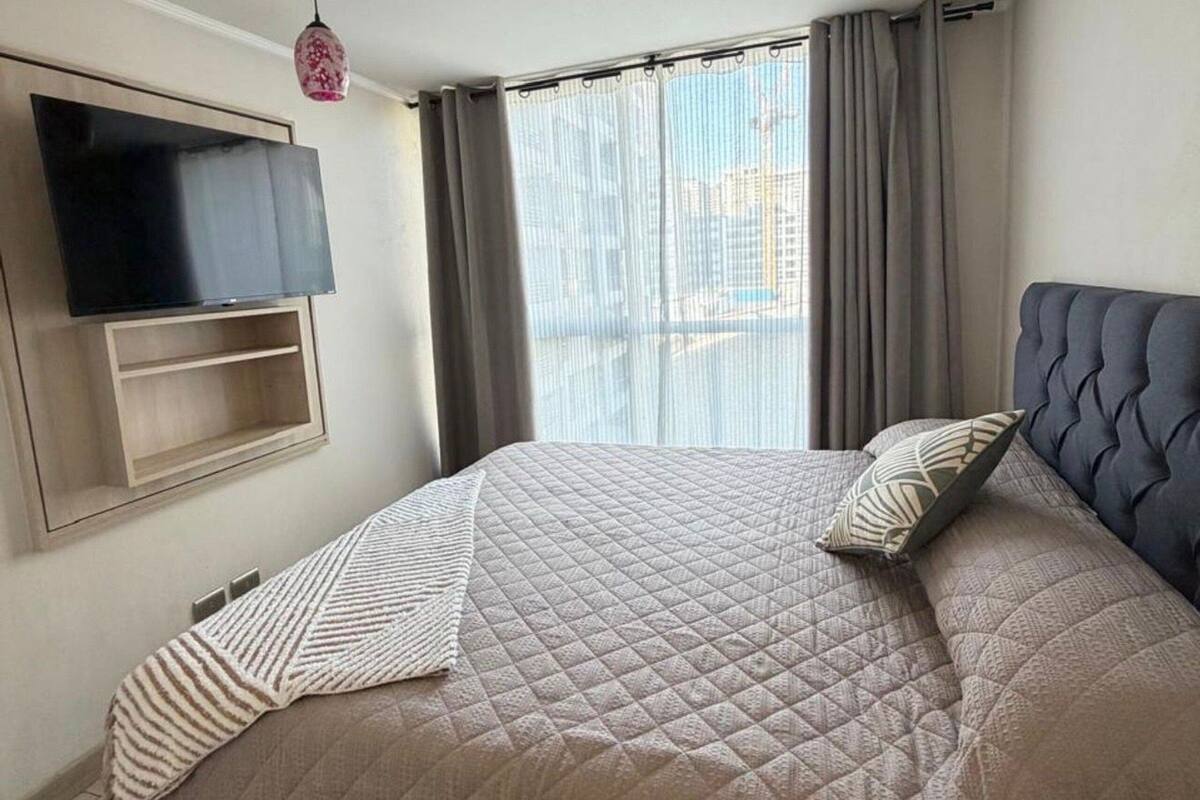 A modern bedroom with a large bed, a mounted TV, and a view of the city through sheer curtains.