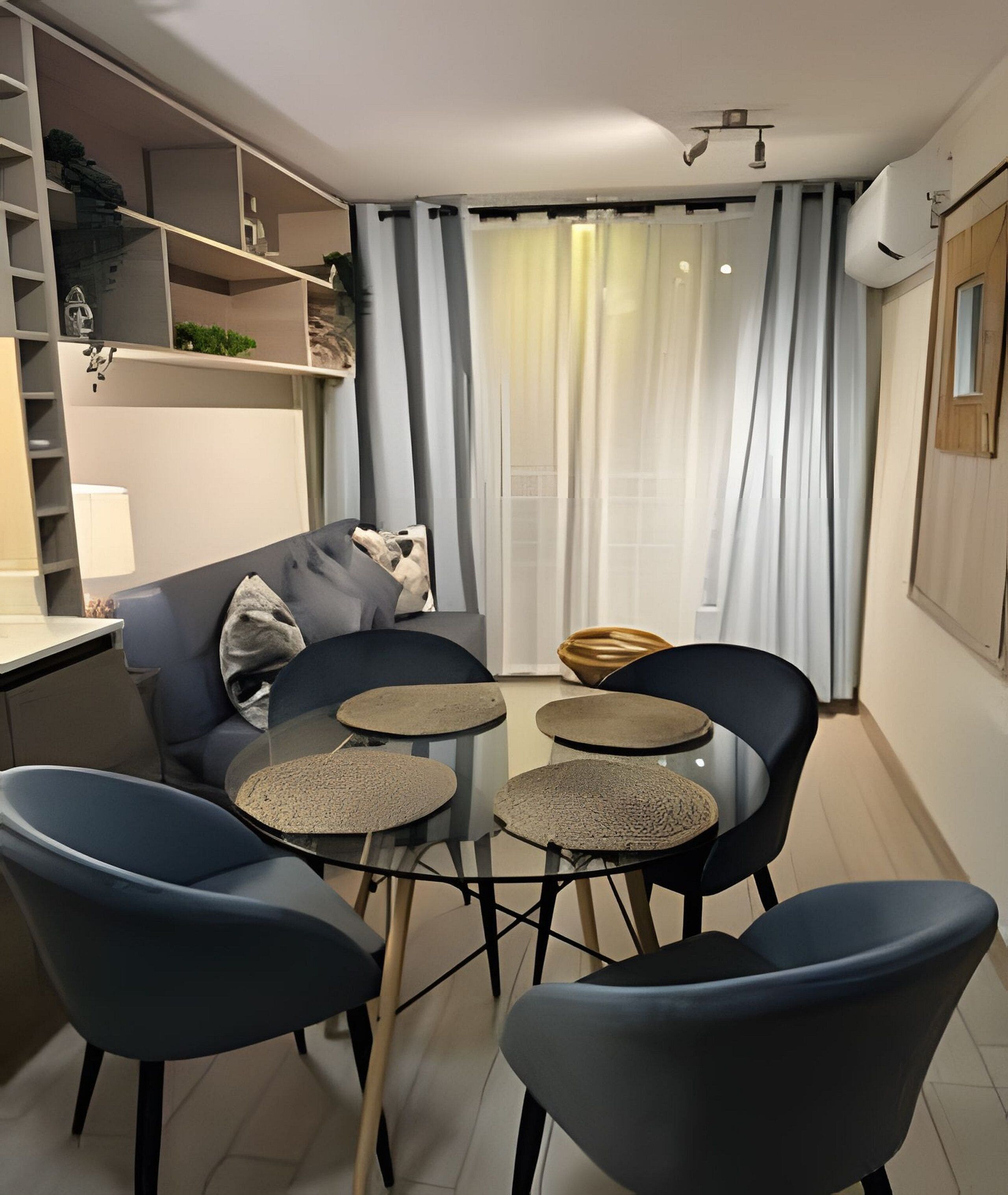 Un salon moderne avec un canapé, deux fauteuils et une petite table ronde. On y trouve une fenêtre avec des rideaux, une étagère intégrée ornée d’objets décoratifs et un climatiseur fixé au mur.
