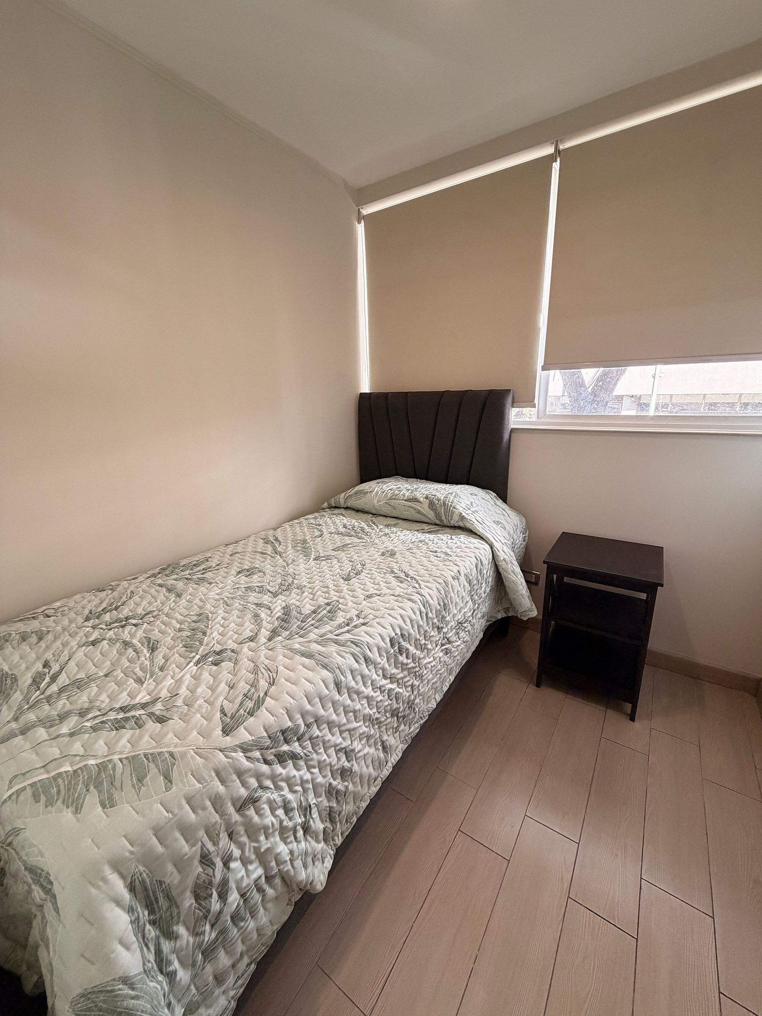 A bedroom with a bed, a nightstand, a window with blinds, and a view of the outside.