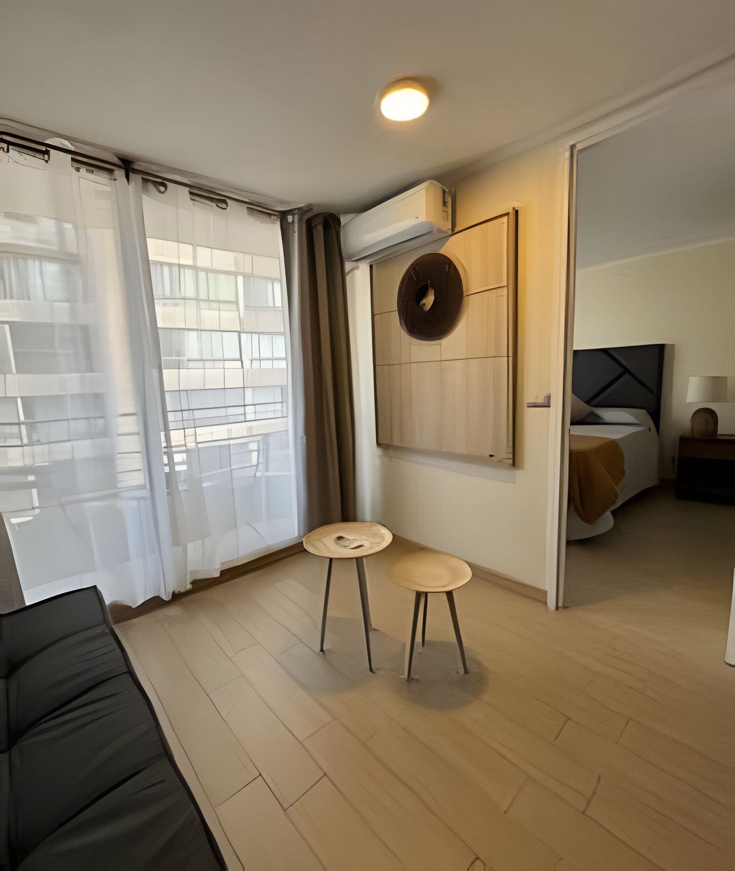 A modern hotel room with a bed, a small table, and a chair. There is a window with curtains, a wall-mounted air conditioner, and a ceiling light.