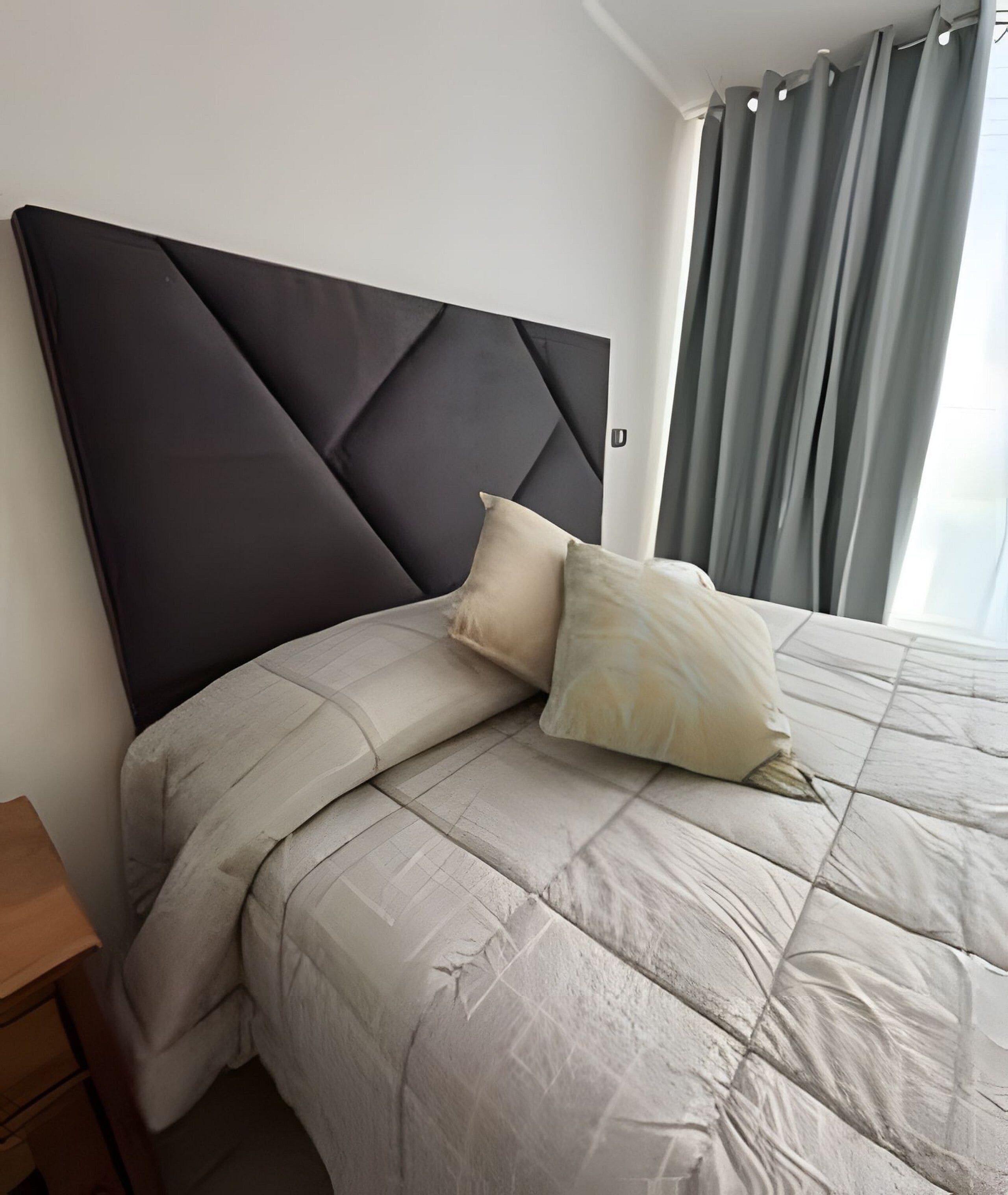 A neatly made bed with a dark headboard and two pillows, a wooden nightstand to the left, and a window with curtains to the right.