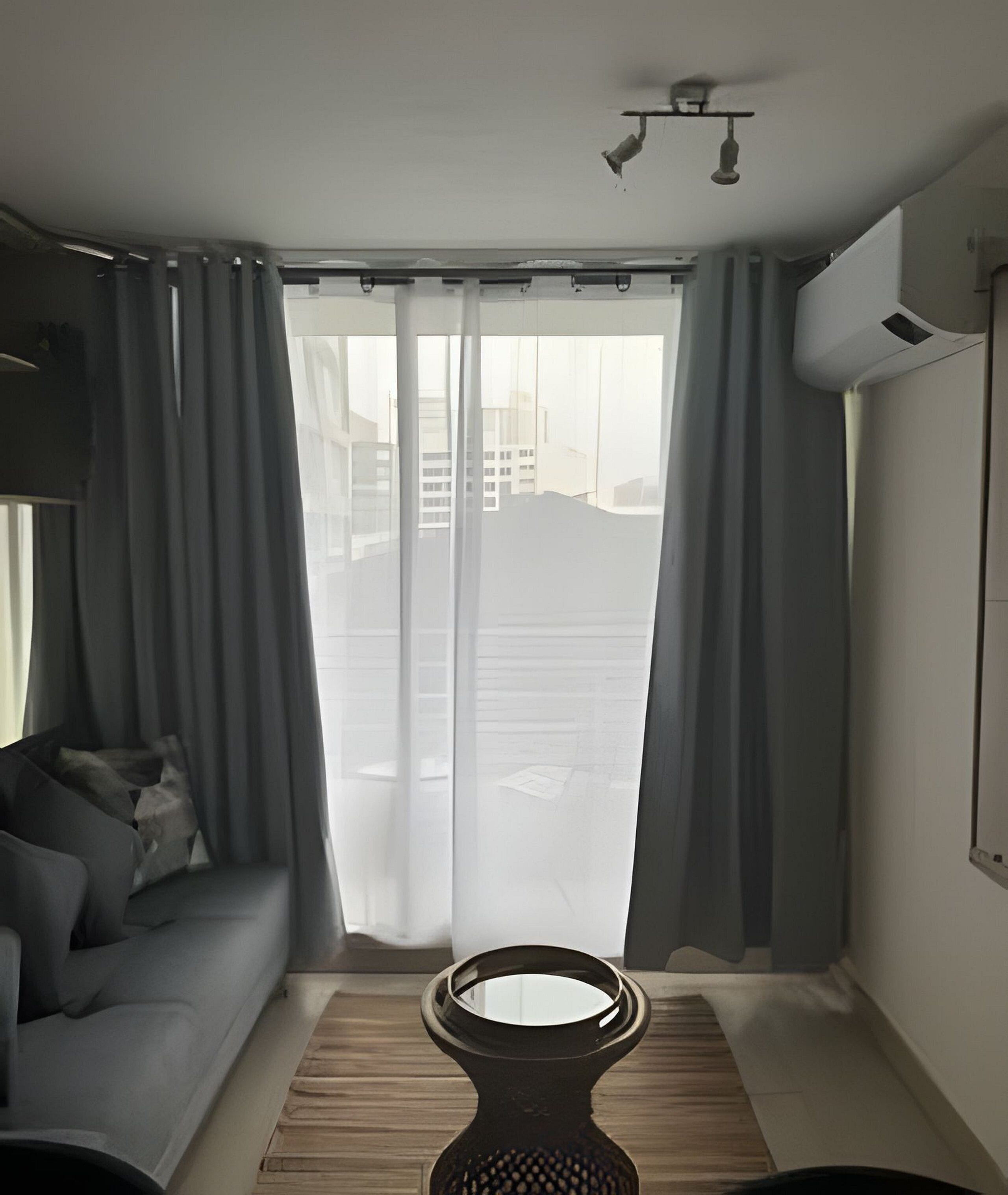Ett modernt vardagsrum med en grå soffa, ett litet runt bord och utsikt genom ett stort fönster med grå gardiner.