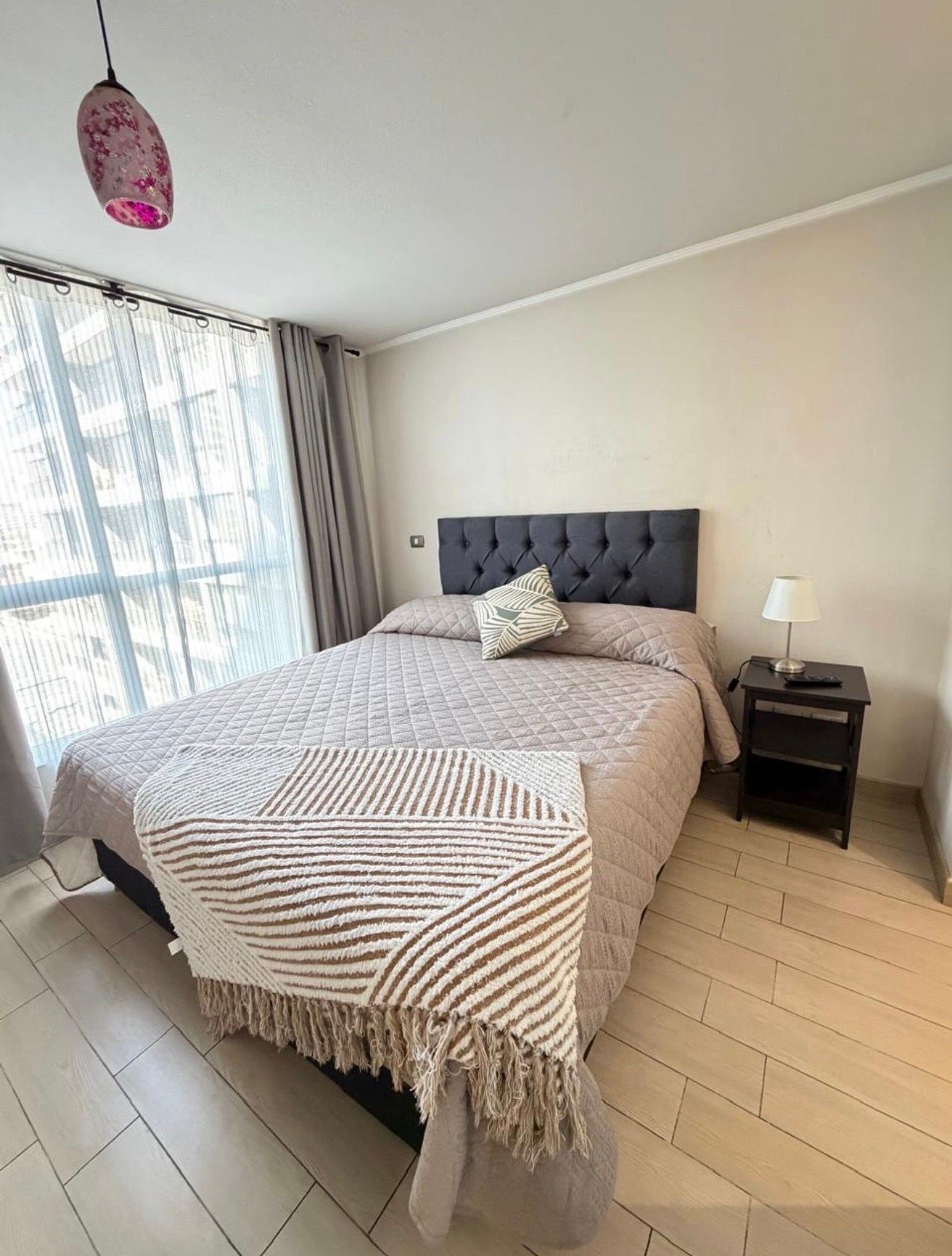 Quarto moderno com cabeceira estofada, uma cama grande com manta listrada, mesa de cabeceira com abajur e vista da cidade através de cortinas finas.
