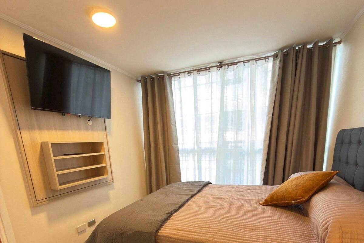 A bedroom with a bed, a mounted TV, a wooden shelf, and a window with curtains.