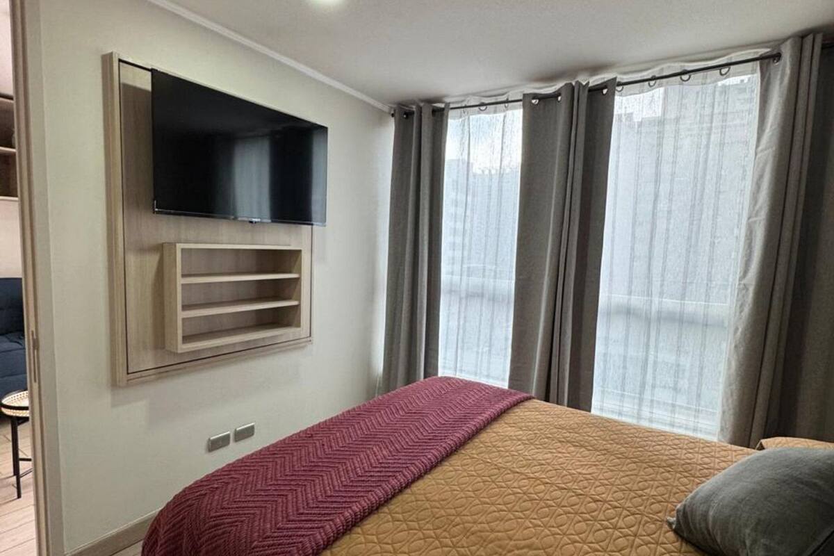 A hotel room with a bed, a mounted TV, a window with curtains, and a wooden floor.