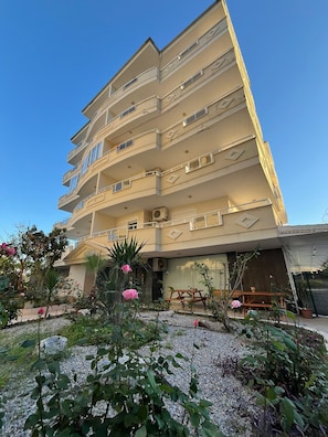 Un edificio de varios pisos con balcones, un área de jardín con plantas y flores, y un patio con mobiliario exterior.